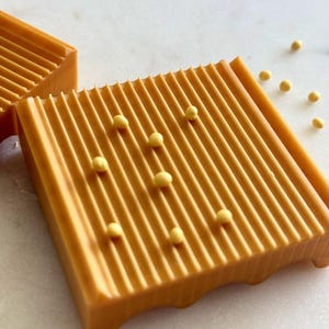 May include: Close-up of two rectangular, orange-colored bars with parallel ridges. Small, yellow spherical objects are scattered around the bars. The bars have a smooth, glossy finish and are set against a white background.