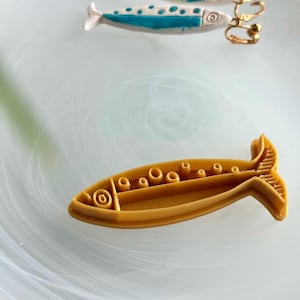 Puede incluir: Un par de pendientes de clip y un cortador de galletas con forma de pez. Los pendientes son blancos con detalles azules y clips dorados. El cortador de galletas es de color amarillo mostaza.