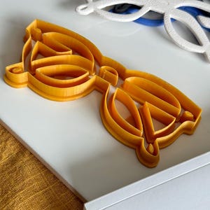 Puede incluir: Un cortador de galletas de plástico amarillo con forma de gafas.