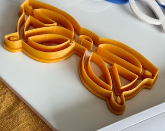 Może przedstawiać: Żółta plastikowa foremka do ciasteczek w kształcie okularów.
