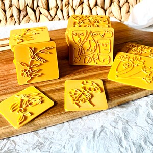 May include: A set of yellow rubber stamps with various designs, including abstract faces, leaves, and flowers. The stamps are arranged on a wooden surface.
