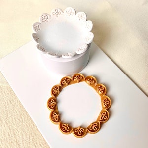 May include: A white ceramic cake decorating mould with a scalloped edge and a delicate floral design. A gold-coloured plastic cake decorating mould with a similar scalloped edge and floral design.