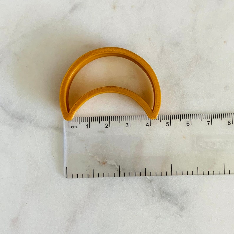 May include: A gold-colored plastic cookie cutter shaped like a crescent moon. The cutter is 4 cm wide and is measured against a ruler.