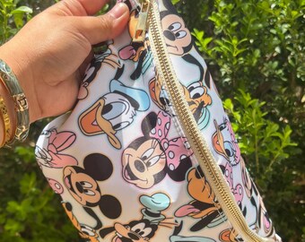 May include: A white fanny pack featuring a colorful all-over print of various cartoon characters including Mickey Mouse, Minnie Mouse, Donald Duck, and Goofy. The bag has a gold-toned zipper closure and an adjustable cream-colored fabric strap.