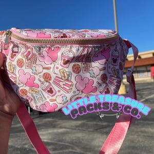 May include: Pink fanny pack with a white background and a Mickey Mouse theme. The pack features a variety of Mickey Mouse ears, snacks, and drinks. The text "Mickey's" is printed on the pack.