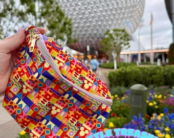 May include: A bum bag featuring a vibrant, multicoloured geometric pattern with small yellow mouse head silhouettes, a gold-tone metal zip, and a light purple adjustable strap. The text Little Treasure Packs & Co. is printed in light blue.
