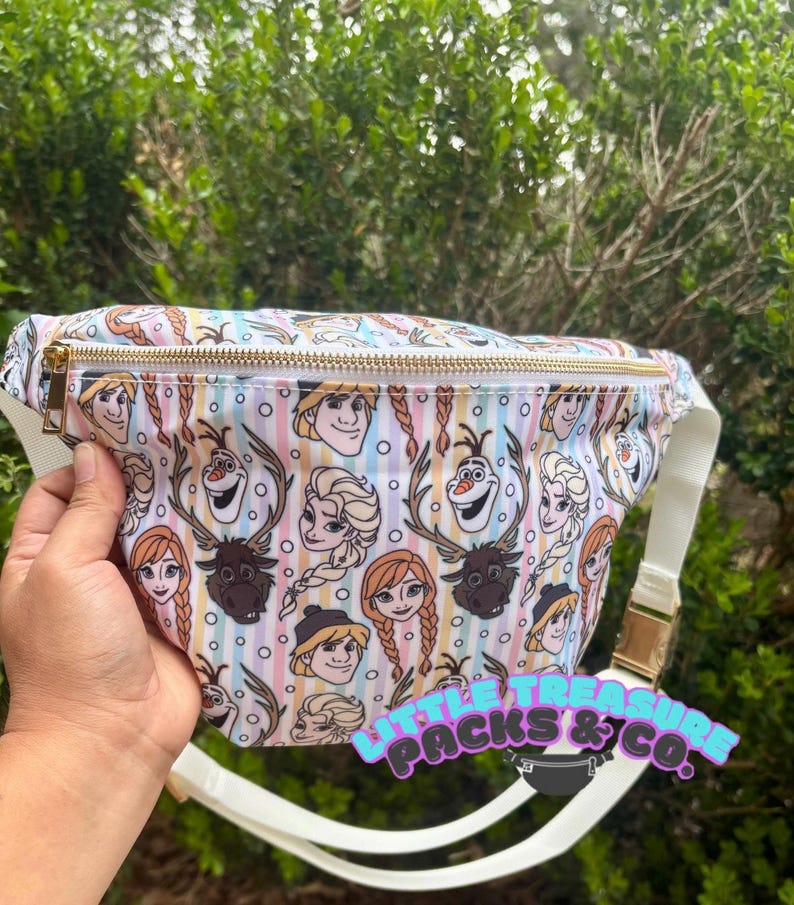 K&ouml;nnte beinhalten: Eine wei&szlig;e Bauchtasche mit goldenem Rei&szlig;verschluss und Schnalle. Die Tasche ist mit Zeichentrickfiguren aus dem Film Frozen, darunter Anna, Elsa, Olaf und Kristoff, gemustert. Die Tasche hat einen wei&szlig;en Riemen und den Text "LITTLE TREASURE PACKS & CO."