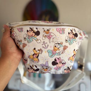 May include: A white fanny pack featuring a repeating pattern of Disney characters including Mickey Mouse, Minnie Mouse, Donald Duck, Daisy Duck, and Pluto dressed in nautical attire, accented with anchors, stars, and a gold-toned zipper closure.