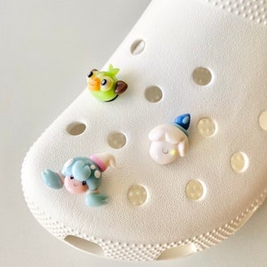May include: Three colourful cartoon charms attached to a white shoe. The charms are a green bird, a white sheep with a blue hat, and a blue crab with pink and white details.