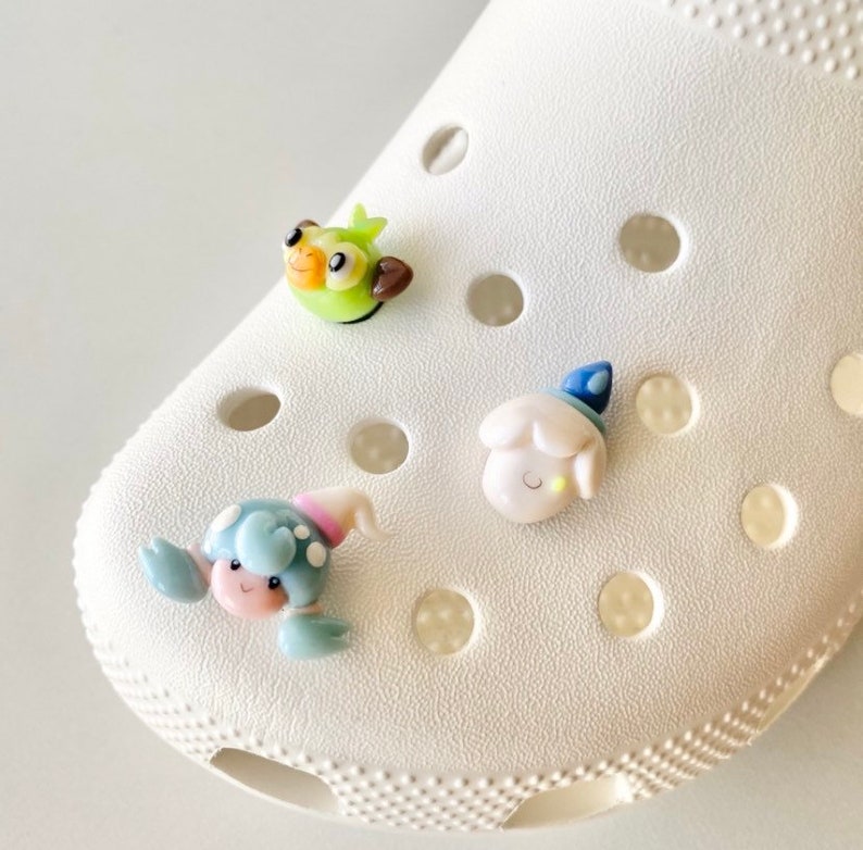 May include: Three colourful cartoon charms attached to a white shoe. The charms are a green bird, a white sheep with a blue hat, and a blue crab with pink and white details.