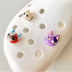 May include: Three colourful animal character charms for a pair of white clogs. The charms are a white and brown dog, a red and blue dog, and a purple cat.