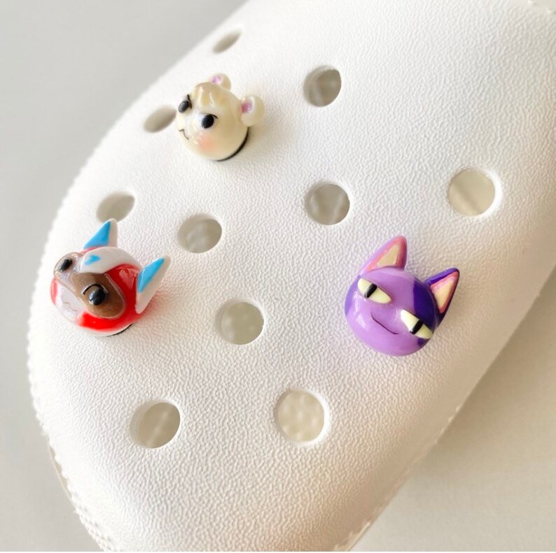 May include: Three colourful animal character charms for a pair of white clogs. The charms are a white and brown dog, a red and blue dog, and a purple cat.