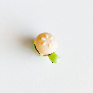 May include: A small, green turtle figurine with a white and yellow hamburger bun on its back. The bun has sesame seeds on top.