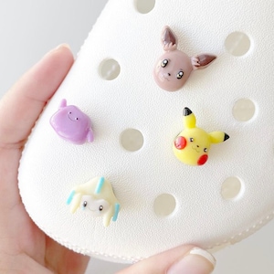 May include: Four colourful cartoon character charms featuring Pokemon characters. The charms are made of plastic and are attached to a white shoe.