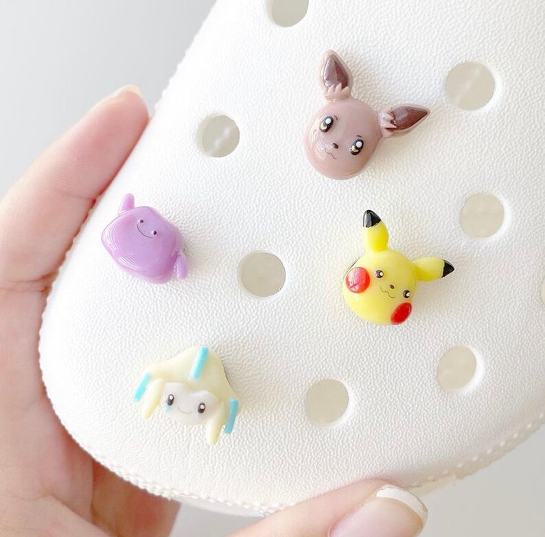 May include: Four colourful cartoon character charms featuring Pokemon characters. The charms are made of plastic and are attached to a white shoe.