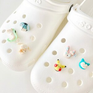 May include: White clogs with colourful cartoon character charms attached. The charms include a blue and green bug, a white rabbit, a pink and blue rabbit, a yellow Pikachu, and a blue cat.