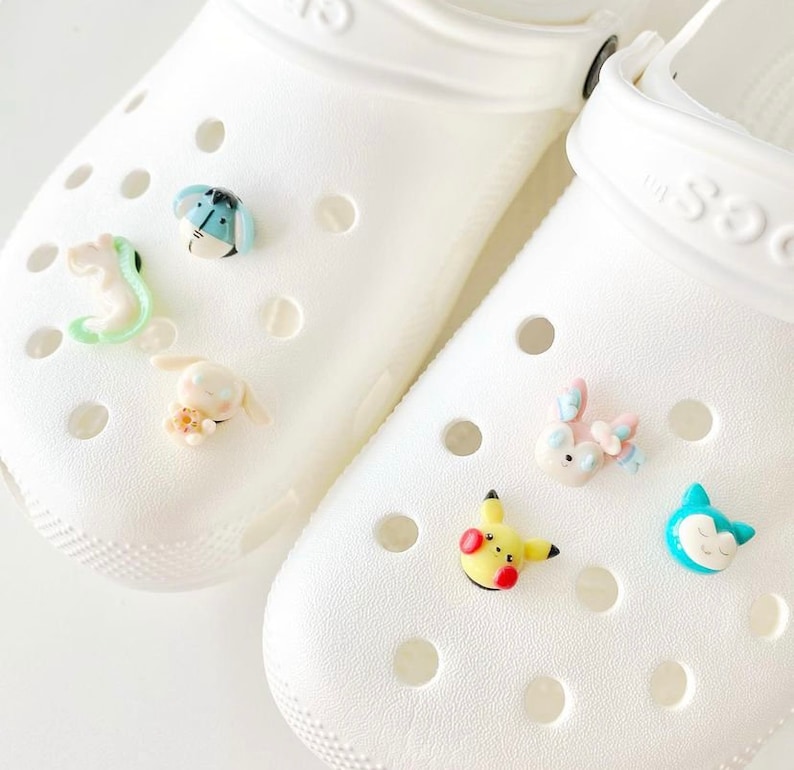 May include: White clogs with colourful cartoon character charms attached. The charms include a blue and green bug, a white rabbit, a pink and blue rabbit, a yellow Pikachu, and a blue cat.