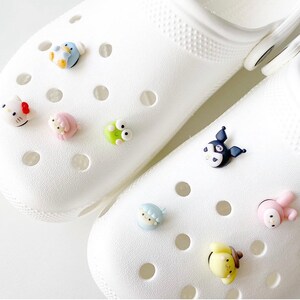 May include: Two white clogs with various colourful cartoon character shoe charms. The charms include a penguin, a frog, a Hello Kitty-like character, a bunny, a bear, and a black and white character with a bow.