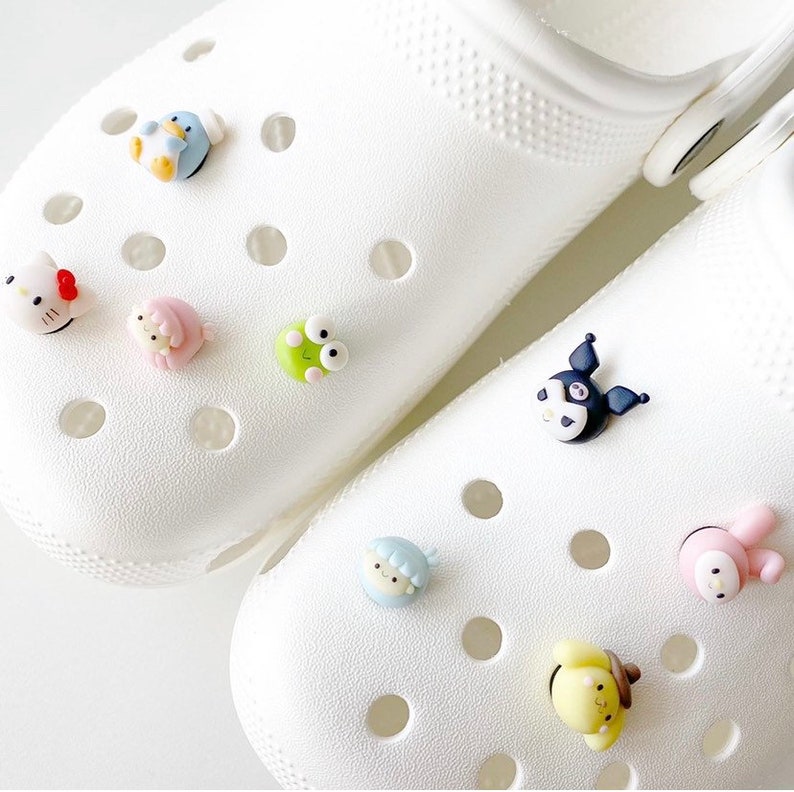 May include: Two white clogs with various colourful cartoon character shoe charms. The charms include a penguin, a frog, a Hello Kitty-like character, a bunny, a bear, and a black and white character with a bow.