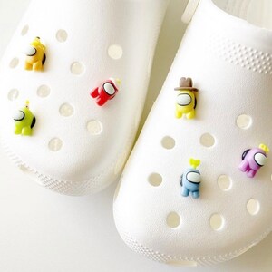May include: White clogs with colourful Among Us character shoe charms. The charms are red, yellow, green, blue and purple.