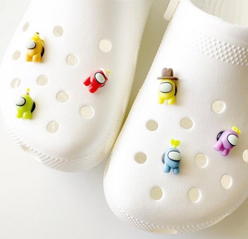 May include: White clogs with colourful Among Us character shoe charms. The charms are red, yellow, green, blue and purple.