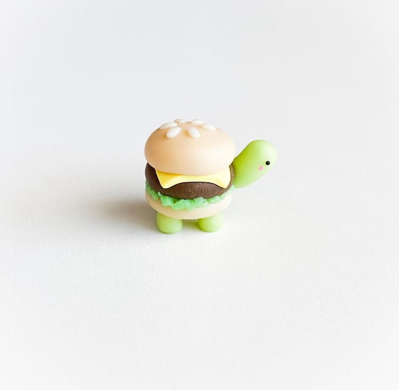 May include: A small, green turtle figurine with a hamburger on its back. The hamburger is made of fondant and has a sesame seed bun, a beef patty, cheese, and lettuce.