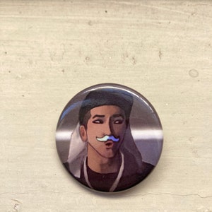May include: A round button with a portrait of a person with a light blue mustache. The person is wearing a dark jacket and a black hat. The background is a gradient of gray and white.