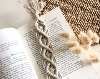 Peut inclure: Un marque-page en macramé beige avec un pompon. Le marque-page est posé sur un livre ouvert avec du texte en français.