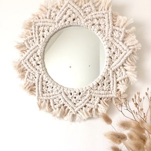 Peut inclure: Un miroir en macram&eacute; blanc avec un miroir circulaire au centre et une bordure en franges. Le miroir est suspendu &agrave; un mur blanc.