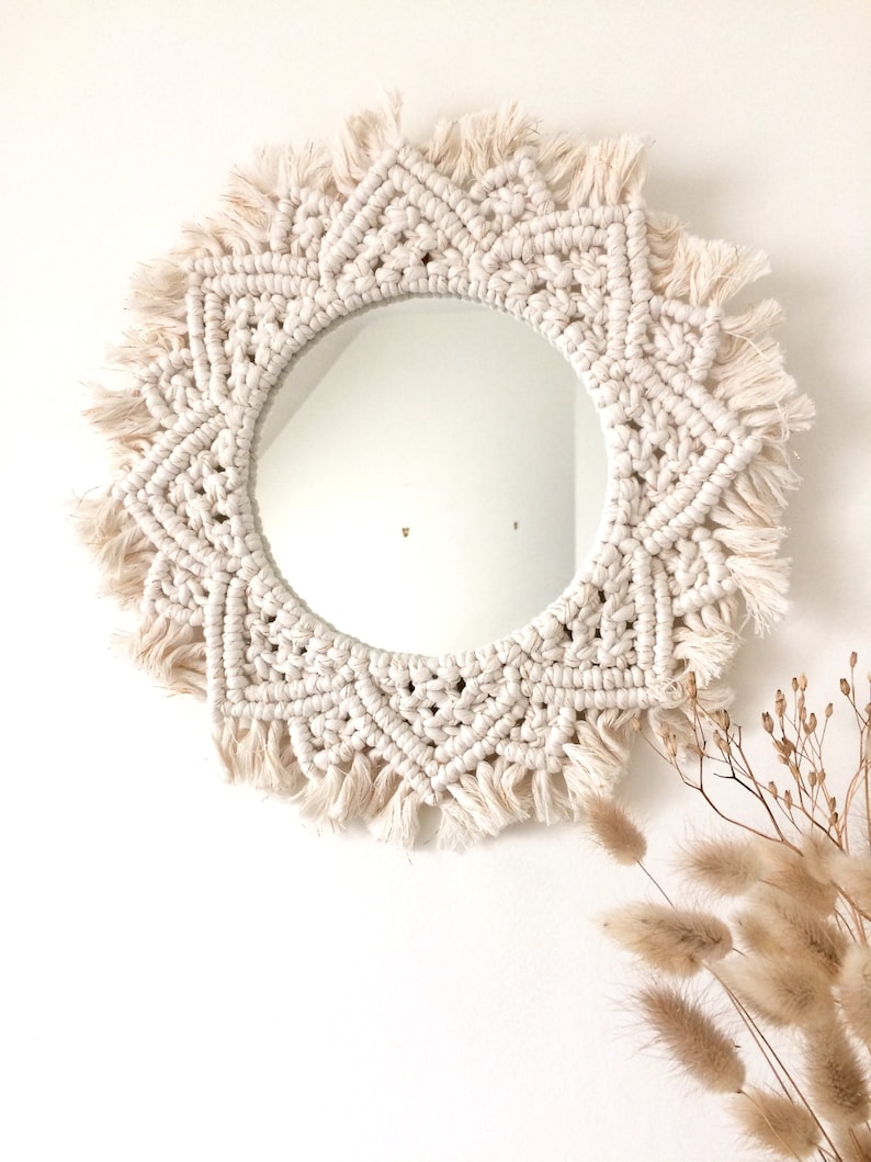 Peut inclure: Un miroir en macram&eacute; blanc avec un miroir circulaire au centre et une bordure en franges. Le miroir est suspendu &agrave; un mur blanc.