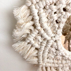 Peut inclure: Un miroir en macram&eacute; blanc avec des accents de fil dor&eacute; et des franges. Le miroir est rond et a un fond blanc.