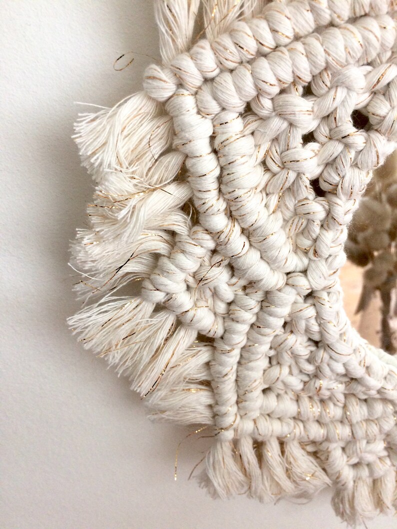 Peut inclure: Un miroir en macram&eacute; blanc avec des accents de fil dor&eacute; et des franges. Le miroir est rond et a un fond blanc.