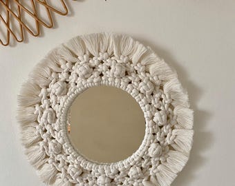 Peut inclure: Miroir rond en macramé blanc cassé, avec une surface réfléchissante centrale. Le design en macramé présente des détails noués et une bordure à franges. Un petit miroir en rotin tressé est visible en haut à gauche.