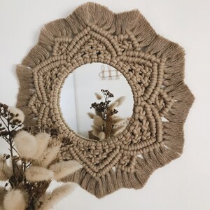 Peut inclure: Un miroir rond avec un cadre en macram&eacute; de couleur beige naturel. Le cadre a une bordure en frange et un motif g&eacute;om&eacute;trique. Le miroir refl&egrave;te un bouquet de fleurs s&eacute;ch&eacute;es.
