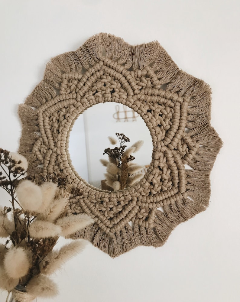 Peut inclure: Un miroir rond avec un cadre en macram&eacute; de couleur beige naturel. Le cadre a une bordure en frange et un motif g&eacute;om&eacute;trique. Le miroir refl&egrave;te un bouquet de fleurs s&eacute;ch&eacute;es.