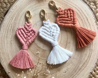 Peut inclure: Trois porte-clés en macramé en forme de cœur avec des pompons. Les porte-clés sont dans des tons de rose, blanc et orange. Les porte-clés sont présentés sur une surface en bois.