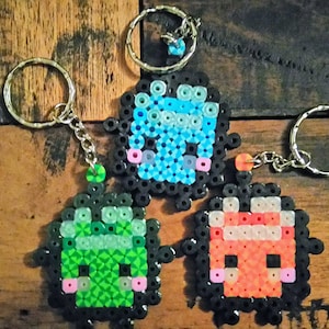 May include: Three pixel art keychains with black bead borders. The keychain on the left is green with pink cheeks, the middle keychain is blue with pink cheeks, and the keychain on the right is orange with pink cheeks.
