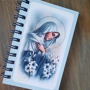 May include: A light blue spiral-bound notebook featuring a rectangular illustration of a figure wearing a blue head covering and robe, with hands crossed over the chest, surrounded by white lily flowers and a thin circular halo line above.