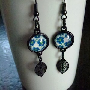Cabochonörhängen med blå blommor på vit bakgrund, små svarta metallbladberlocker, monterade på svarta metallstolpar och krokar