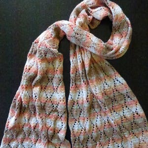 Könnte beinhalten: Gestrickter Schal in Pfirsich-, Braun- und Cremetönen mit einem rautenförmigen Lochmuster. Der Schal liegt auf einer dunklen Oberfläche, wodurch sein filigranes Design und seine weiche Textur hervorgehoben werden. Ein elegantes Accessoire.