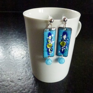 Op de afbeelding: Een paar rechthoekige hangende oorbellen met een felblauwe emaille afwerking, een donkerblauwe middenstreep, witte spikkels en een kleine limoengroene accentkraal. Elke oorbel eindigt onderaan met een ronde, lichtblauwe kraal.