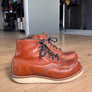 Könnte beinhalten: Ein Paar knöchelhohe Stiefel aus glattem, rotbraunem Leder. Die Stiefel verfügen über eine Mokassin-Zehenpartie mit markanten weißen Kontrastnähten, silberfarbene Metallösen und schwarz-weiß gesprenkelte Schnürsenkel auf einer dicken, cremefarbenen Sohle.