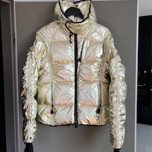 Puede incluir: Una chaqueta acolchada dorada metálica con ribetes negros y cuello alto. La chaqueta tiene cierre de cremallera y varios bolsillos.