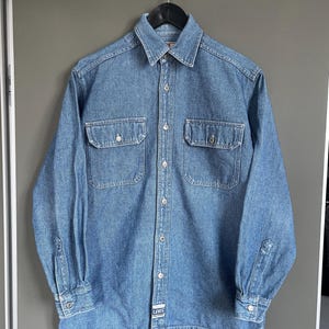 May include: A light blue denim shirt with long sleeves, two chest pockets, and button closures. The shirt is hanging on a black hanger against a gray background. The Levi's logo is visible on the bottom.