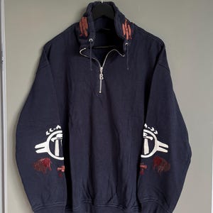 May include: A dark blue sweatshirt with a half-zip closure and white embroidered designs on the sleeves. The designs feature a stylized image of a hammer and a bison. The collar has red embroidered details.