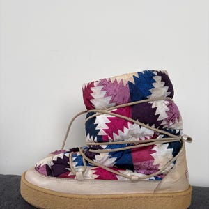 May include: A colourful winter boot featuring a geometric pattern in pink, purple, blue, and white. The boot has a beige sole and a lace-up design. The boot's height is approximately 20 cm.