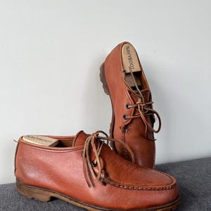May include: A pair of brown leather shoes with laces. The shoes have a moccasin-style toe and a sturdy sole. Shoe trees are inserted in the shoes. The shoes are displayed on a grey surface against a white background.