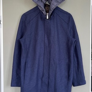 May include: A navy blue coat with a detachable hood. The coat has a zipper closure and a tailored fit. The hood is a lighter shade of blue and has a quilted design. The coat is hanging on a door.