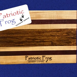 May include: A wooden cutting board with a patriotic theme. The board is made of maple, walnut, and purpleheart wood. It has a natural finish and is engraved with the words "Patriotic Frog" and "Veteran Owned".