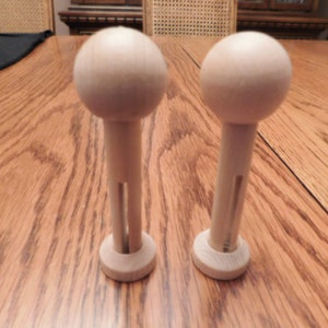May include: Two unfinished wooden doll pins with a rounded top and a cylindrical stem. The pins have a small circular base and a slot cut into the stem. The items are made of light-colored wood and are displayed on a wooden surface.